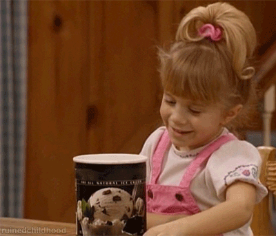 A young girl sitting at a table, her eyes sparkling with joy as she swirls her spoon and digs in to a carton of the ultimate tempting food: ice cream.