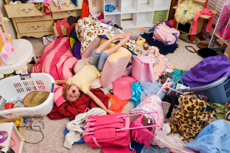 A child lying upon a pile of clothes and toys in a chaotic messy bedroom